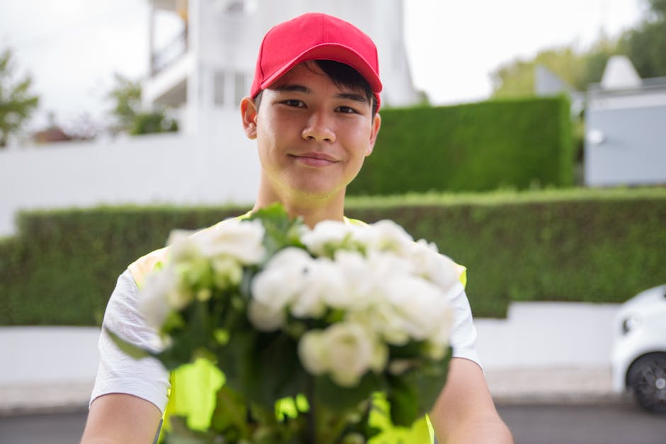A smiling young man wearing a red cap and a yellow reflective vest is holding out a bouquet of fresh white flowers, including roses and ranunculus, wrapped in clear cellophane. The bouquet features vi