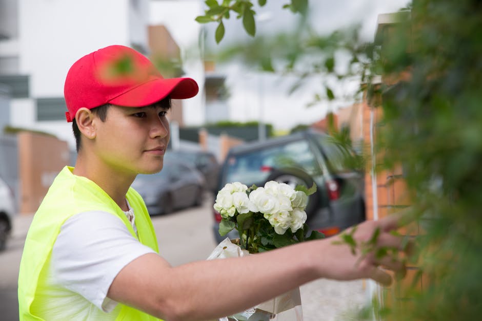 A young delivery person wearing a red cap and a high-visibility yellow vest is arranging a bouquet of white hydrangeas wrapped in white paper, standing outdoors on a residential street. The flowers ar