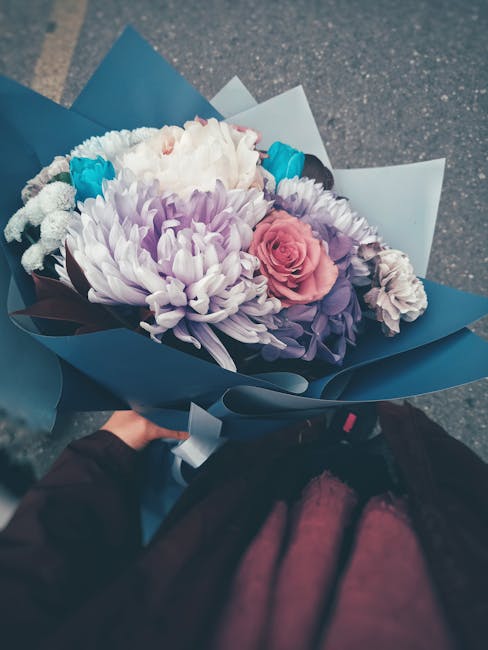 A hand holding a bouquet of fresh flowers wrapped in dark blue paper, featuring a large white chrysanthemum, a soft pink rose, light purple lisianthus, and small blue accents, arranged in a floral gif