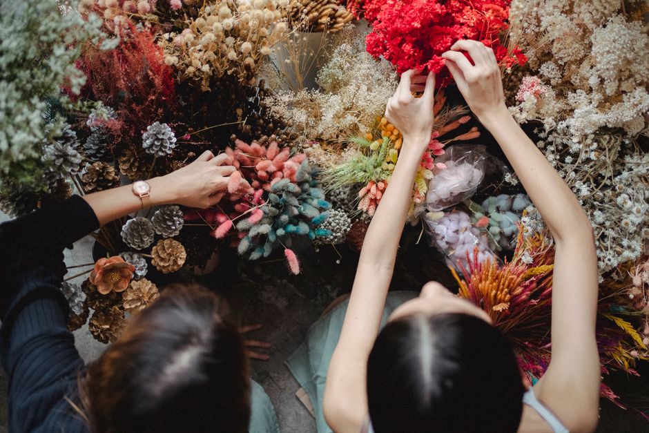 Two florists are seen from above arranging a variety of fresh flowers on a dark surface. The flowers include red, pink, white, and purple blooms, with some dried elements like pinecones and textured b