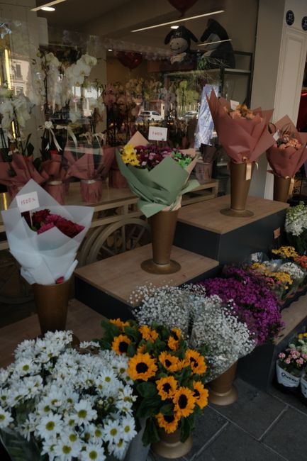 A flower shop display featuring a variety of fresh floral arrangements and bouquets. In the foreground, there are white daisies, yellow sunflowers, and clusters of small purple, white, and pink flower